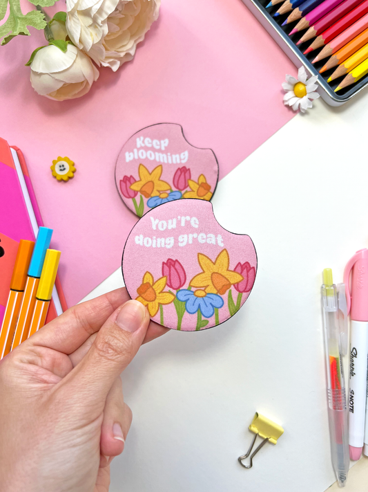 Set of two round floral car coasters with pastel designs and uplifting phrases “Keep Blooming” and “You’re Doing Great,” surrounded by soft flowers.