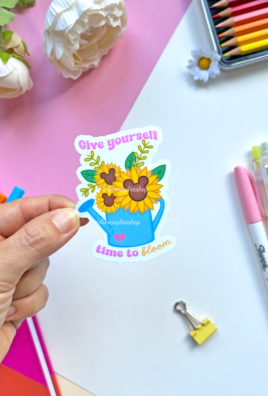 Vinyl sticker of a watering can filled with blooming sunflowers and the message “Give Yourself Time To Bloom” — a symbol of growth and self-care.