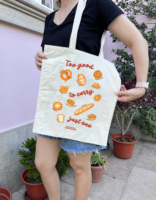 Natural cotton tote bag illustrated with breads and pastries surrounding the phrase "Too Good to Carry Just One".