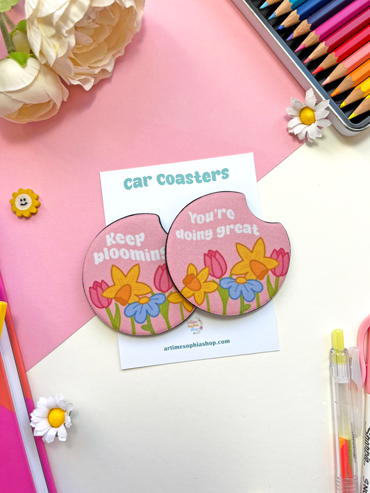 Set of two round floral car coasters with pastel designs and uplifting phrases “Keep Blooming” and “You’re Doing Great,” surrounded by soft flowers.