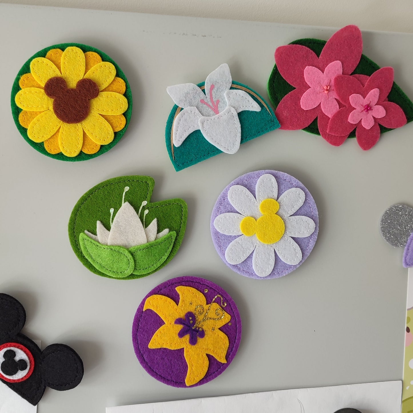 A collection of handmade felt flower accessories inspired by Disney characters — including Mulan’s magnolia, Rapunzel’s sun drop flower, Tiana’s lily pad, a sunflower, and a daisy. Shown styled as keychains, magnets, pins, and hair clips — perfect for Disney Bounds, gifts, or floral decor.